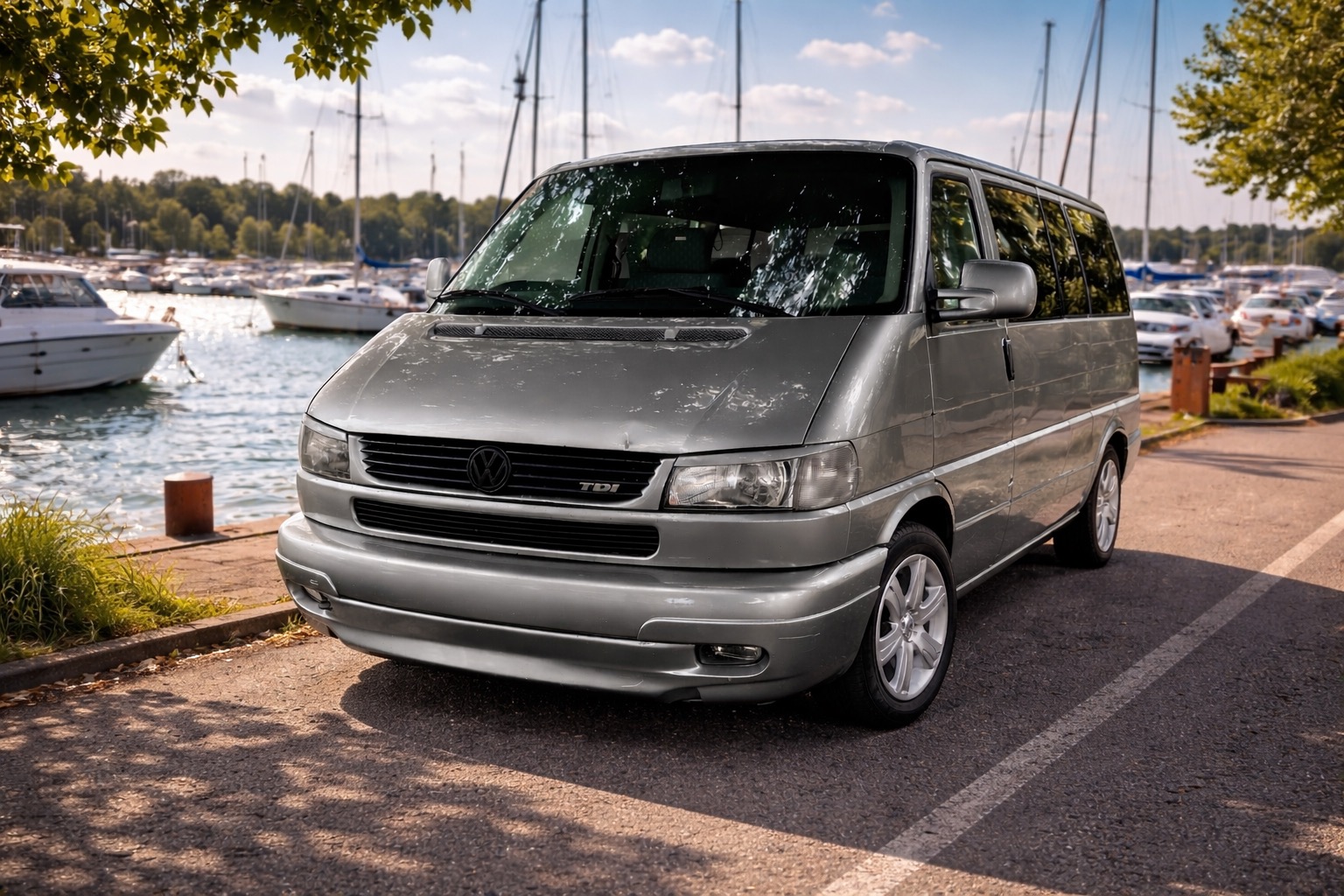 Srebrny bus VW Transporter T4 na tle przystani jachtowej w słoneczny dzień. Widok z przodu, z detalami maski i reflektorów. Idealny do przewozu osób lub transportu.
