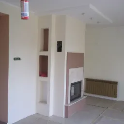 Wnętrze w trakcie prac wykończeniowych: ściany częściowo pomalowane na biało i brązowo, zabudowa kominka z marmurowym elementem, widoczny grzejnik i zabezpieczona podłoga.