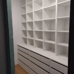 Biała garderoba z szufladami i półkami, widok na wnętrze szafy w trakcie montażu, surowa ściana z płyt gipsowo-kartonowych w tle.