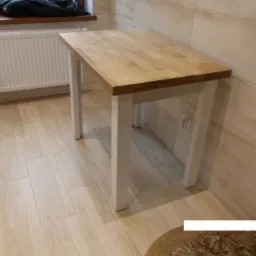 Prostokątny stół z jasnego drewna z białymi nogami, ustawiony w rogu jasnej kuchni, widoczny grzejnik i torba na oknie.