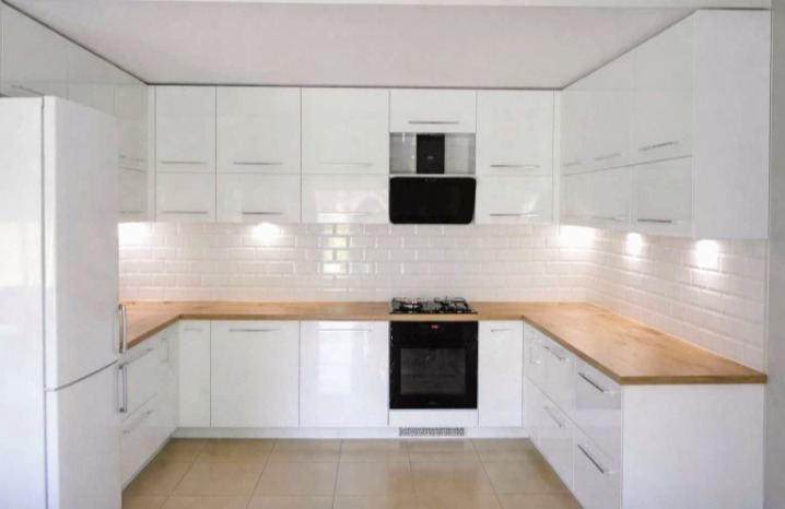 Biała, nowoczesna kuchnia z drewnianym blatem i białymi płytkami na ścianie. Ustawienie w kształcie litery U, oświetlenie punktowe pod szafkami.