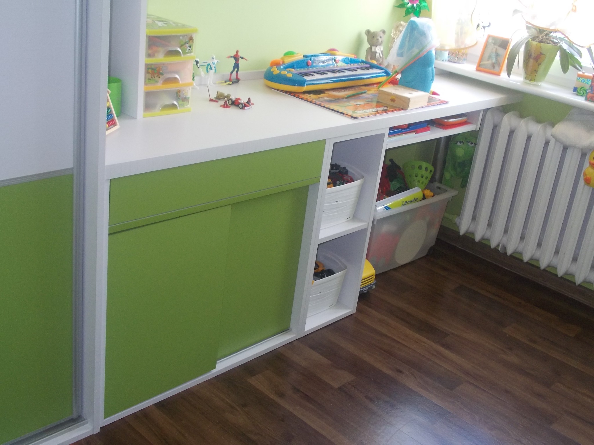Pokój dziecięcy z biurkiem z szafkami i półkami, biały blat, zielone fronty szafek, zabawki na blacie, drewniana podłoga.