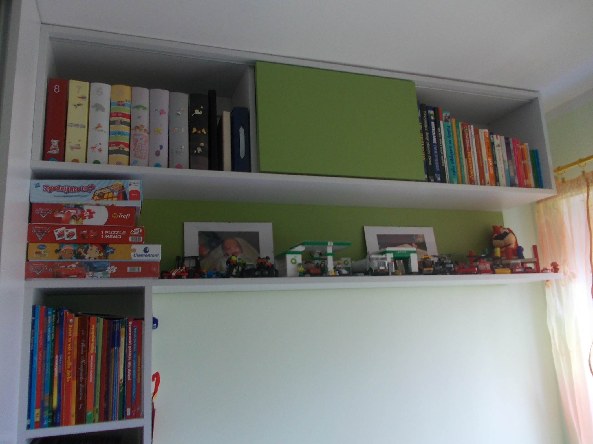 Białe półki w pokoju dziecięcym z książkami, grami planszowymi i kolekcją zabawek Lego, w tym stacje benzynowe, ustawionymi na tle zielonej ściany.
