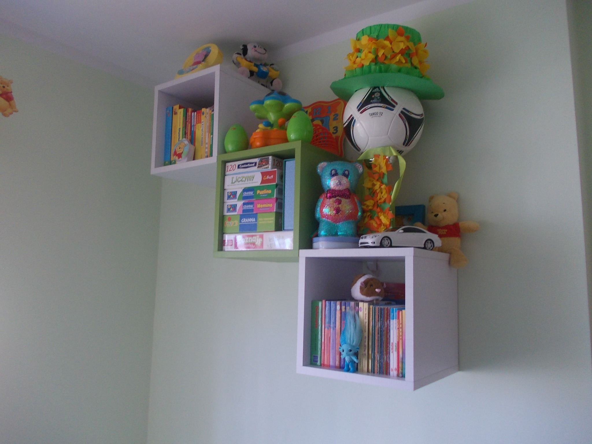 Kaskadowe, białe i zielone półki w pokoju dziecięcym, wypełnione książkami, grami planszowymi, pluszakami i zabawkami, w tym piłką do nogi w zielonym kapeluszu.