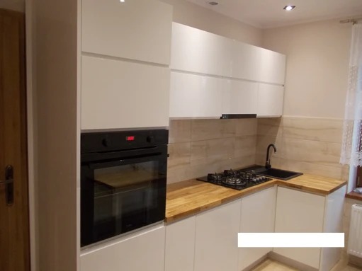 Biała, nowoczesna kuchnia z drewnianym blatem i czarną płytą gazową oraz piekarnikiem w Polanowie, oświetlona punktowymi lampami sufitowymi.