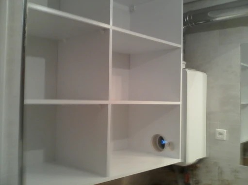 Biała szafka łazienkowa z otwartymi półkami, z widocznym otworem na instalację hydrauliczną i bojlerem na ścianie w tle.