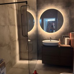 LOGICTOM Tomasz Nowacki - Nowoczesna łazienka z kabiną prysznicową typu walk-in, okrągłym lustrem z podświetleniem LED i umywalką nablatową. Widoczny fragment toalety i narzędzie glazurnicze.