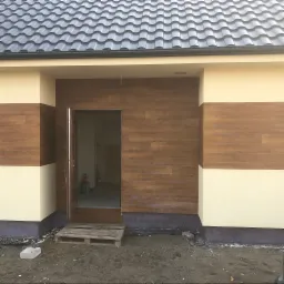 Wejście do budynku z elewacją imitującą drewno, częściowo pomalowaną na kremowo, widoczna paleta i materiały budowlane w otwartych drzwiach.