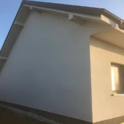 Świeżo otynkowana elewacja domu jednorodzinnego w odcieniach szarości, widok z boku na ścianę z dwoma oknami, widoczne elementy wykończeniowe dachu i fundamentów, w tle zieleń.