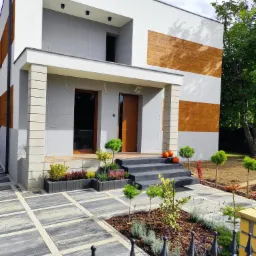 Nowoczesny dom murowany z elementami dekoracyjnymi imitującymi drewno na elewacji, zadbany ogród z kostką brukową i ozdobnymi roślinami, dynie na schodach wejściowych.