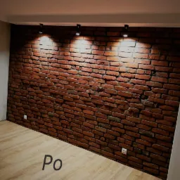 Ściana z dekoracyjnej cegły w pomieszczeniu, oświetlona trzema punktowymi lampami sufitowymi, z podłogą imitującą drewno i widocznym fragmentem napisu 'Po'.