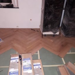 Rafał - Układanie parkietu w jodełkę francuską, widoczne paczki parkietu Creativ, narzędzia, zabezpieczone meble i częściowo ułożona podłoga na zielonym podkładzie.