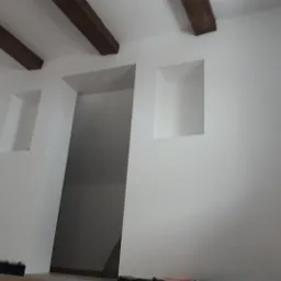 Białe ściany z wnękami i drewnianymi belkami sufitowymi, widok na otwarte przejście, częściowo widoczne listwy przypodłogowe i wiaderko na podłodze.