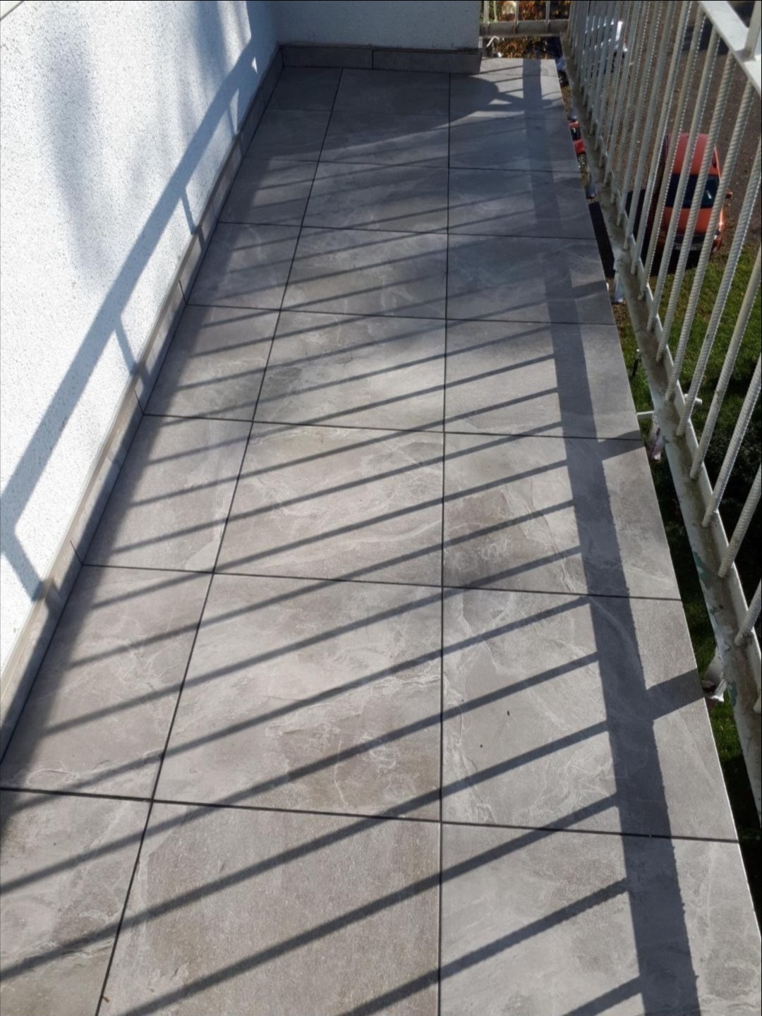 Wyłożony szarymi płytkami balkon z metalową balustradą, cień rzucany przez balustradę, widok z góry.