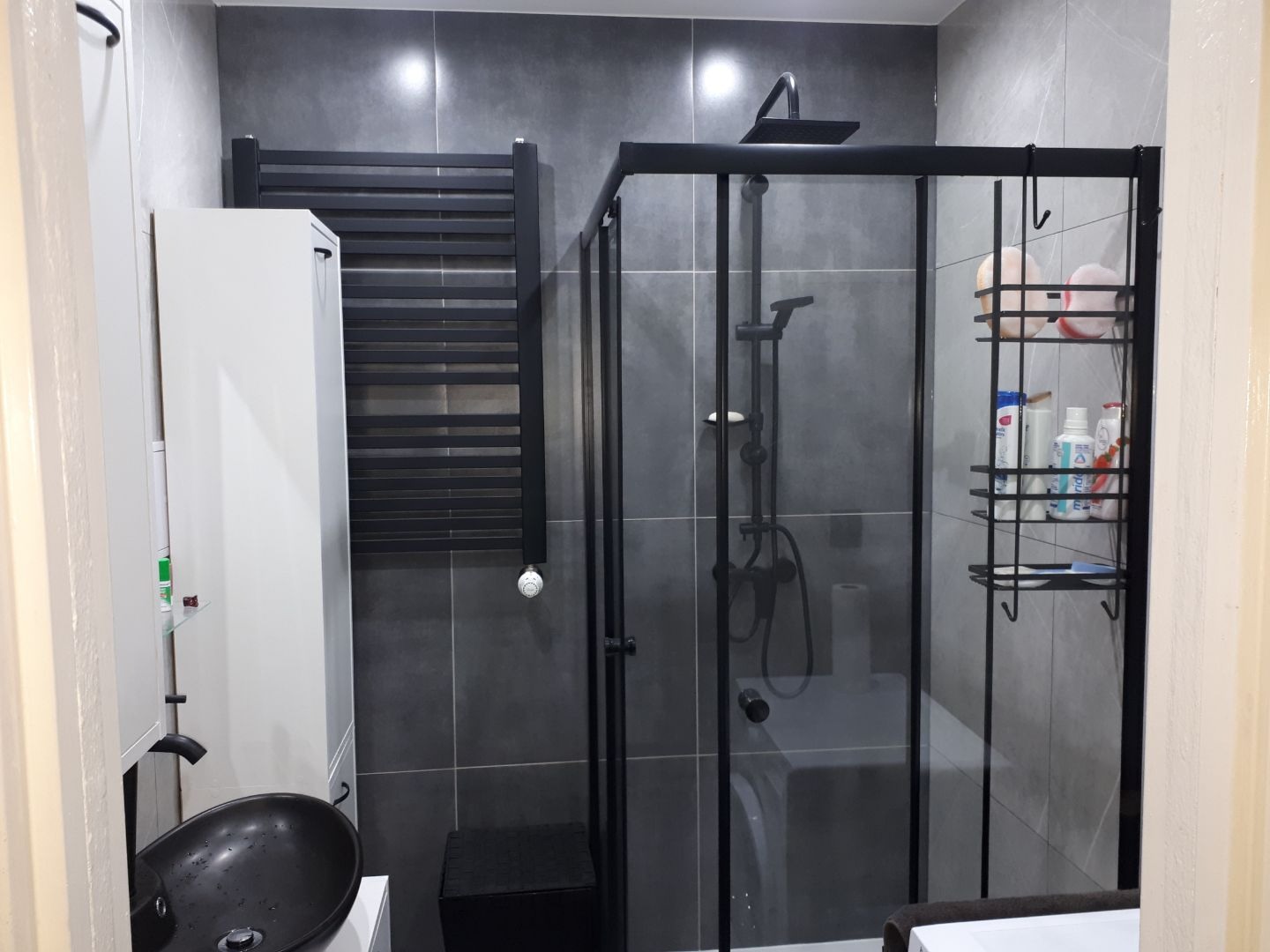 Nowoczesna łazienka z szarymi płytkami, czarną kabiną prysznicową typu walk-in, czarnym grzejnikiem drabinkowym i umywalką nablatową w kształcie misy, z widocznymi kosmetykami na półkach.