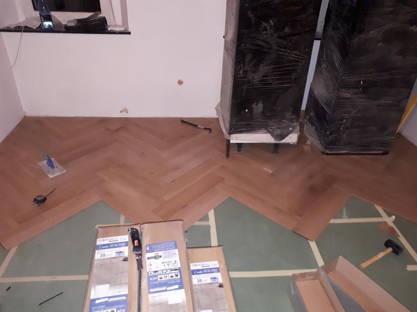 Układanie parkietu w jodełkę francuską, widoczne paczki parkietu Creativ, narzędzia, zabezpieczone meble i częściowo ułożona podłoga na zielonym podkładzie.