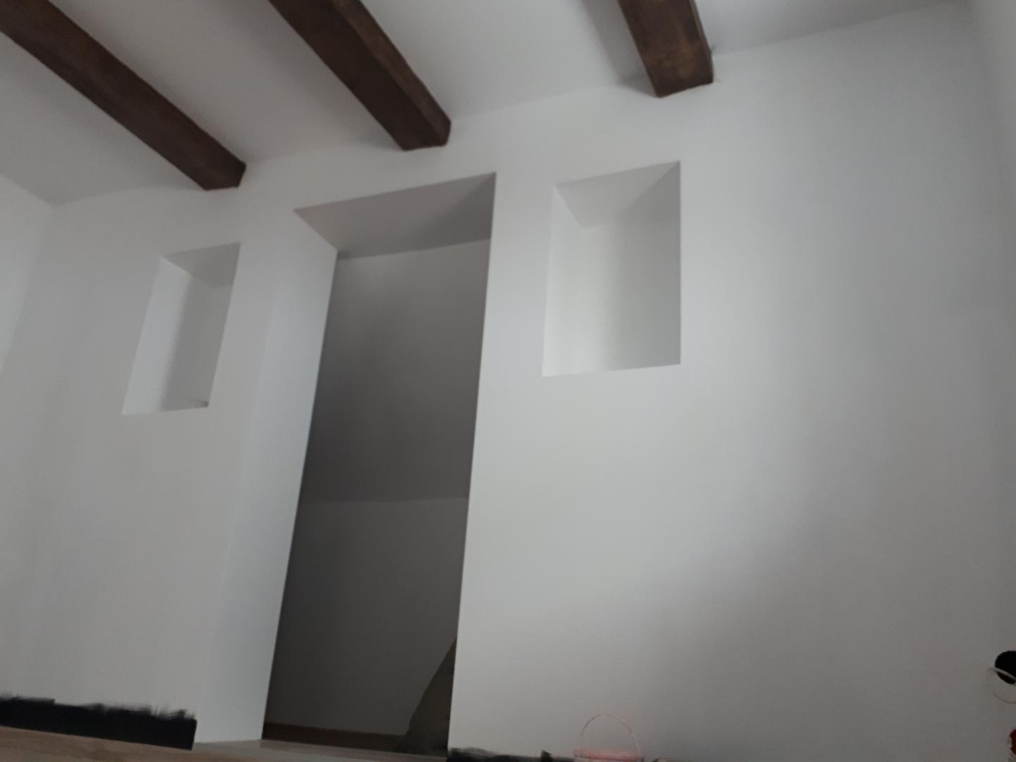 Białe ściany z wnękami i drewnianymi belkami sufitowymi, widok na otwarte przejście, częściowo widoczne listwy przypodłogowe i wiaderko na podłodze.