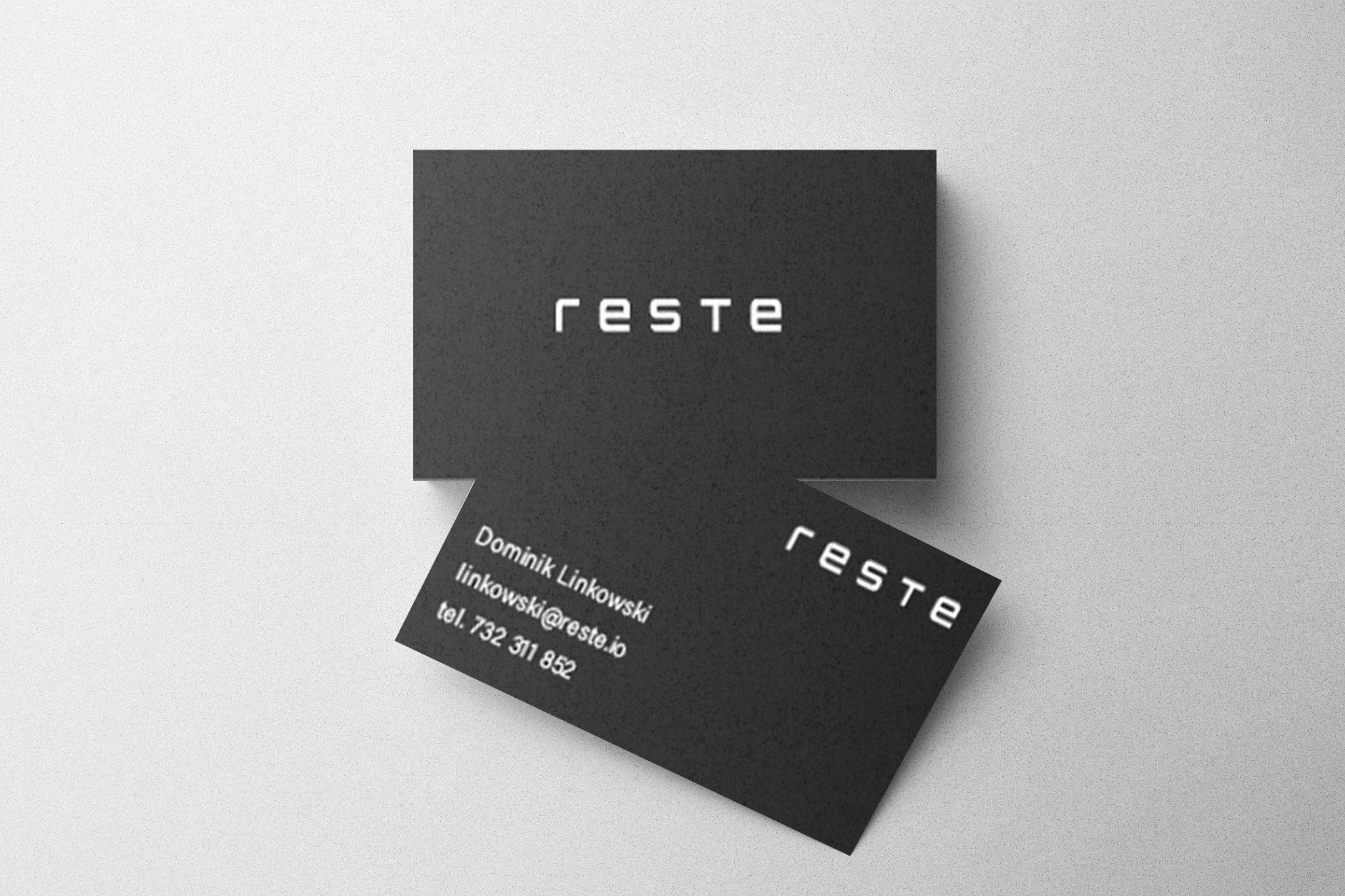 Dwie czarne wizytówki z białym napisem 'reste' oraz danymi kontaktowymi na dolnej wizytówce, leżące na jasnym, teksturowanym tle.