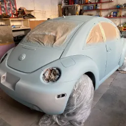 Volkswagen Beetle w trakcie lakierowania, zabezpieczony taśmą i folią, widoczne przygotowania do nałożenia kolejnej warstwy lakieru w warsztacie.