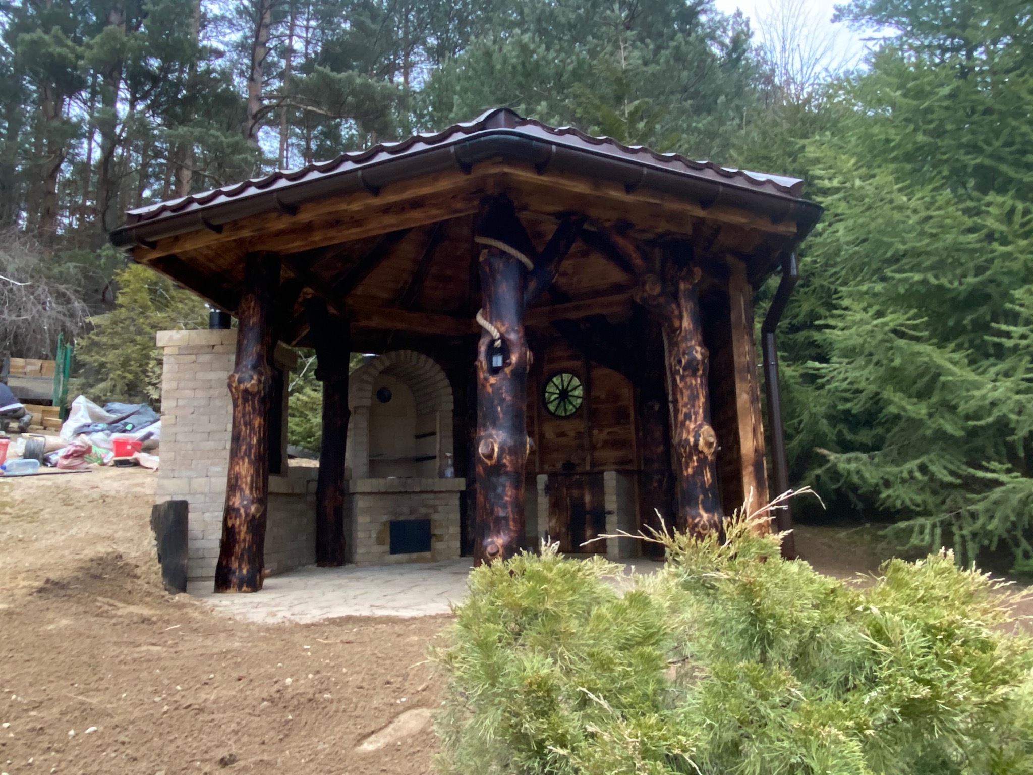 Drewniana altana z kominkiem i wędzarnią wbudowana w skarpę, z elementami z surowego drewna imitującego pnie drzew, okrągłe okienko i ceglany komin.