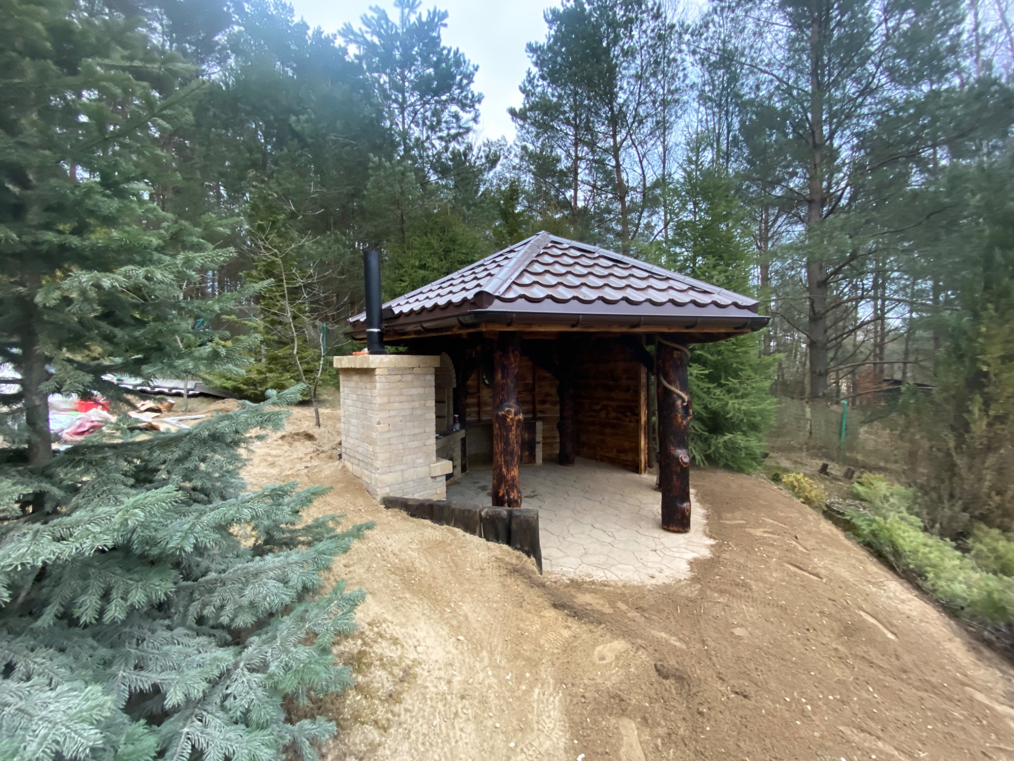 Zadaszona konstrukcja z ceglanym kominem i drewnianymi słupami, prawdopodobnie grillowa, z brązowym dachem i betonową posadzką, otoczona drzewami iglastymi i świeżo wyrównanym terenem.