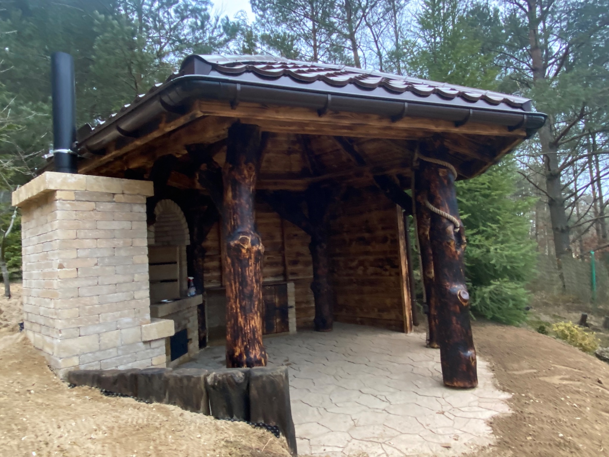 Zadaszona konstrukcja z piecem chlebowym, drewniane słupy imitujące pnie drzew, brązowy dach z rynną, kamienna podłoga, otoczenie leśne.