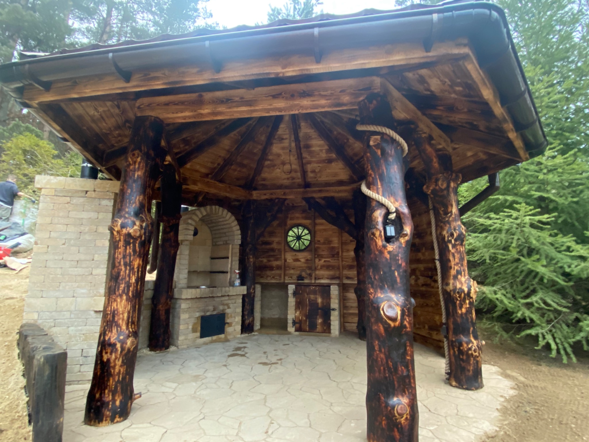 Zadaszona konstrukcja z drewnianymi słupami imitującymi pnie drzew, z grillem murowanym z cegły i drewnianymi drzwiczkami, na kamiennej posadzce, w tle iglaste drzewa.
