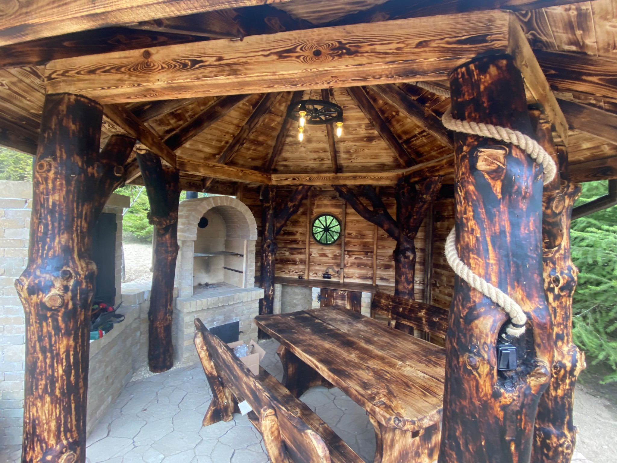Zadaszona konstrukcja z drewna stylizowanego na naturalne pnie drzew, z oświetleniem w stylu retro, murowanym grillem i okrągłym oknem, wyposażona w masywny drewniany stół z ławami.