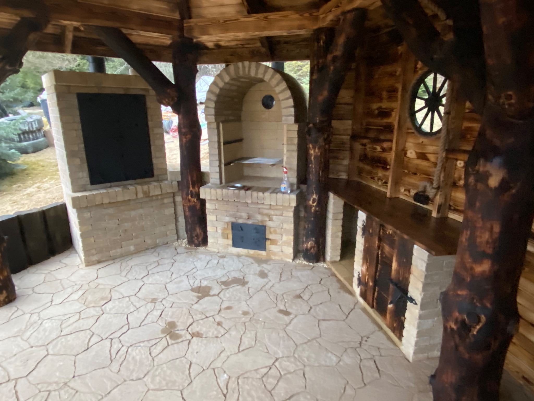 Zadaszona altana z grillem murowanym z cegły i drewnianym blatem roboczym, wykończona elementami z surowego drewna imitującego pnie drzew.