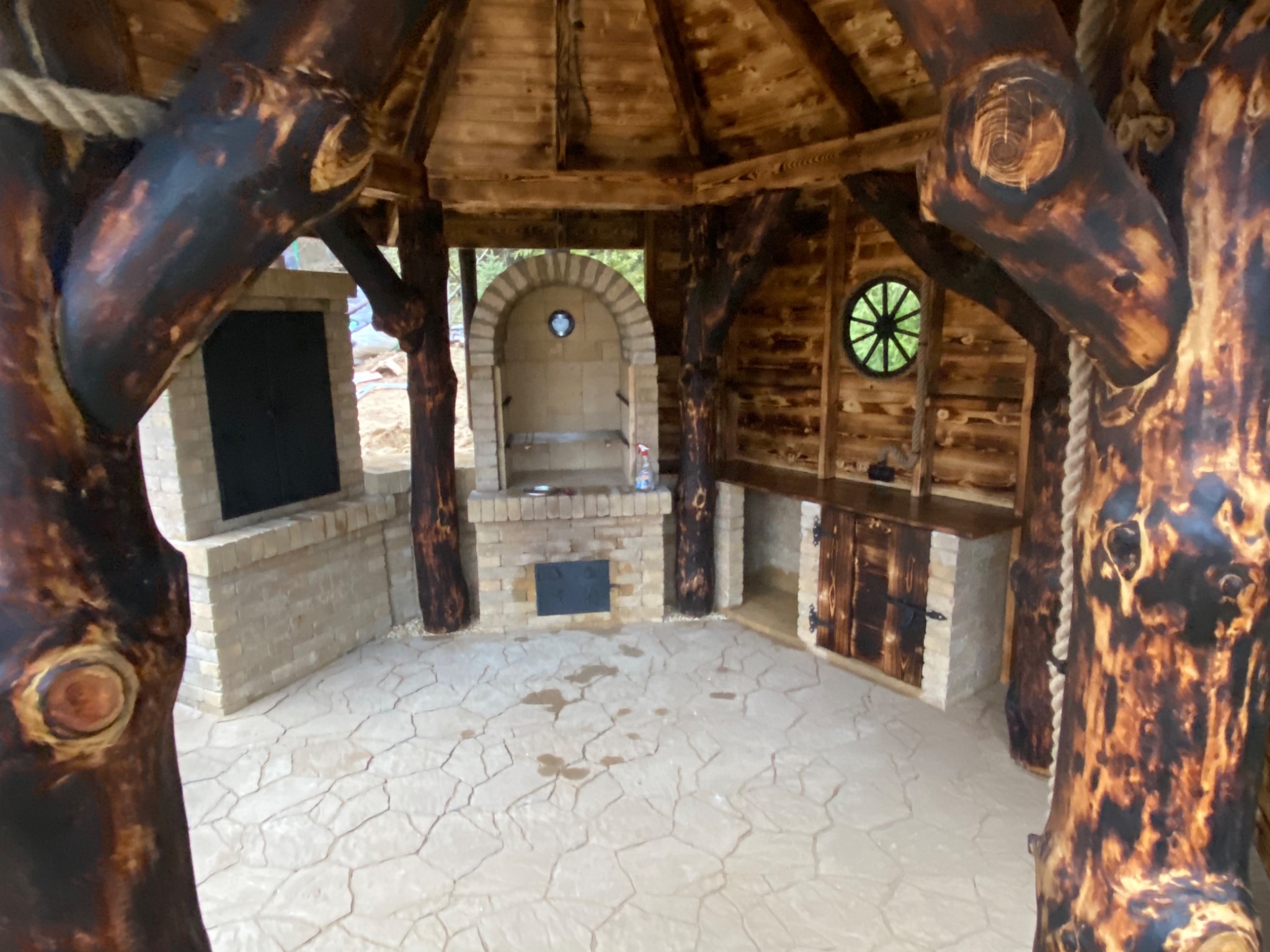 Wnętrze altany z piecem do pizzy i blatem roboczym, drewniane elementy konstrukcyjne z wyraźnym rysunkiem słojów, podłoga wyłożona płytami imitującymi kamień.