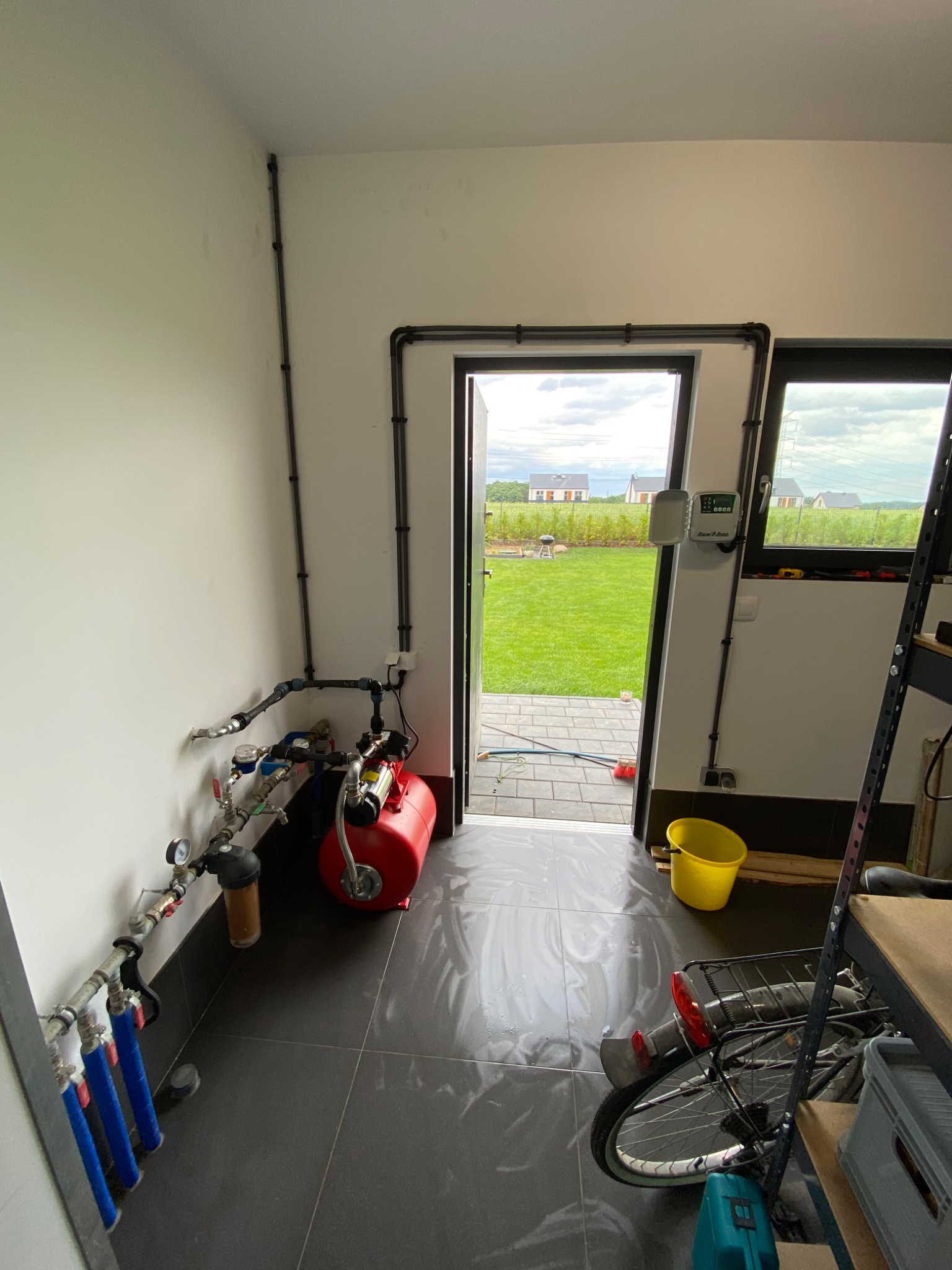 Instalacja systemu nawadniania w pomieszczeniu gospodarczym, widoczny czerwony zbiornik ciśnieniowy, filtry, rury, sterownik Rain Bird oraz widok na trawnik przez otwarte drzwi.