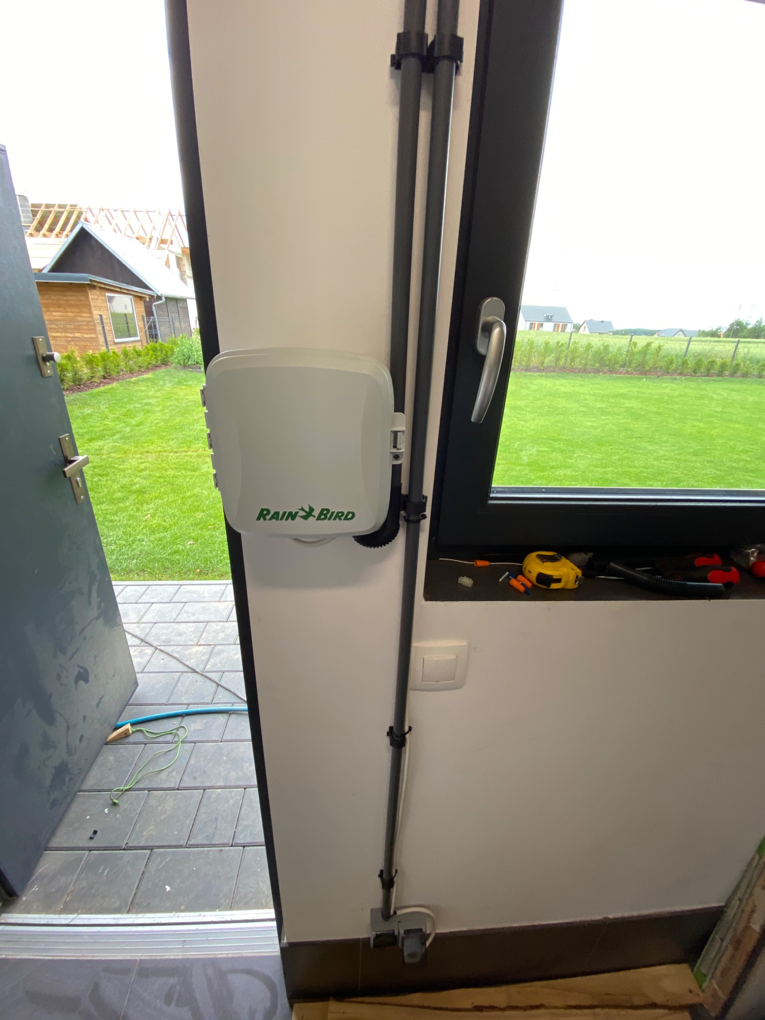 Zamontowany sterownik nawadniania Rain Bird na białej ścianie wewnątrz budynku, widoczne rury doprowadzające wodę, okno z narzędziami i miarą zwiniętą, oraz fragment ogrodu z trawnikiem na zewnątrz.