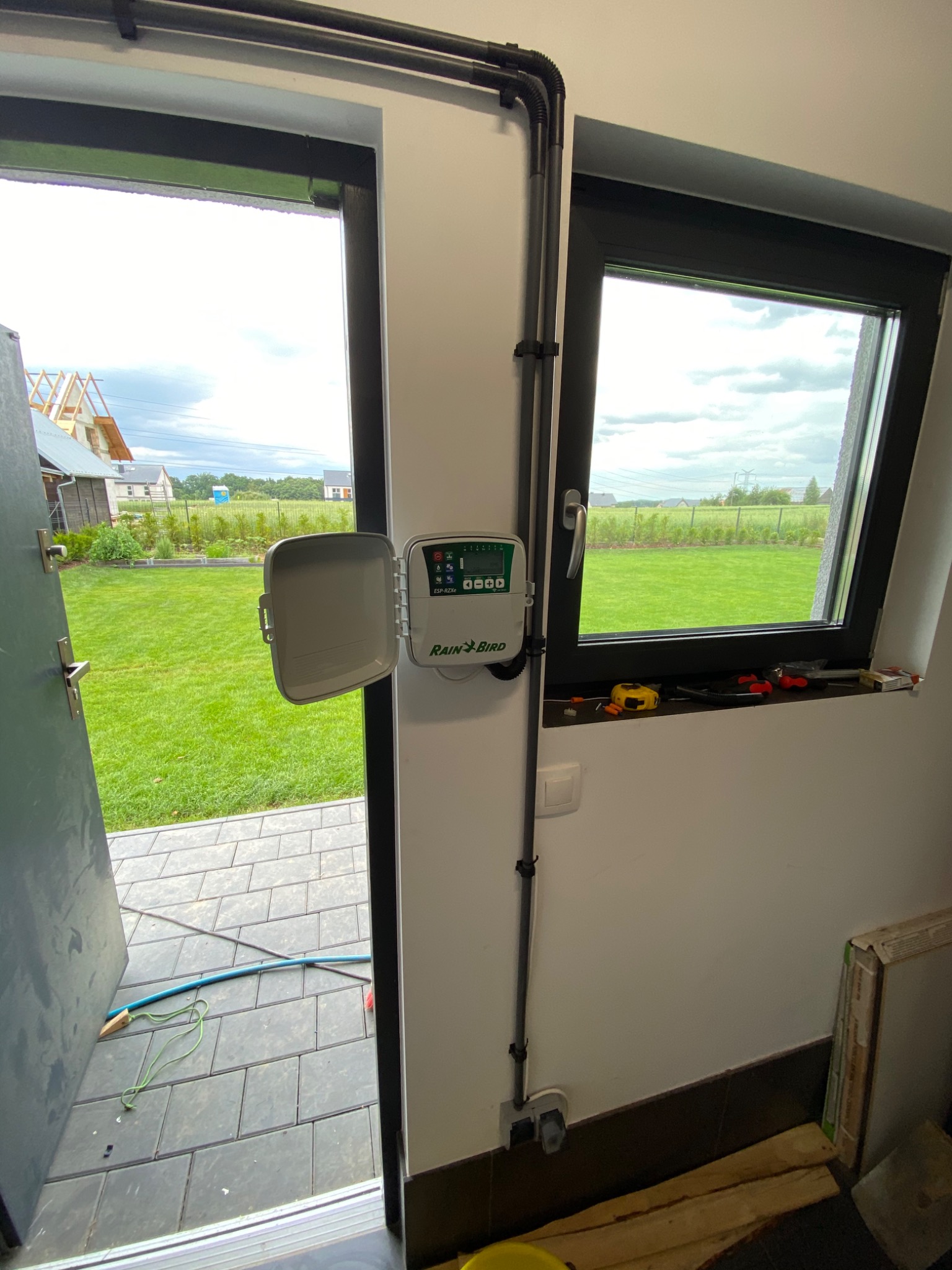Panel sterowania systemu nawadniania Rain Bird zamontowany na ścianie, widok z wnętrza na ogród i dom w budowie przez otwarte drzwi.