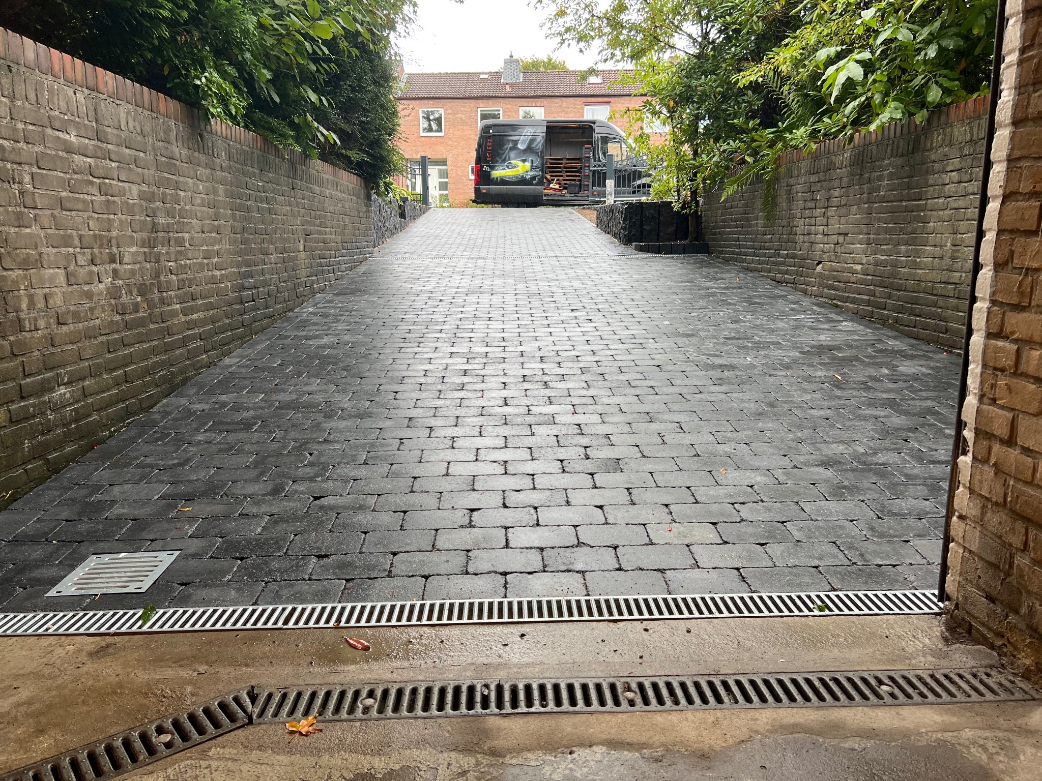 Podjazd z kostki brukowej w kolorze grafitu z systemem odprowadzania wody, zakończony murem z cegły oraz samochodem dostawczym w tle.