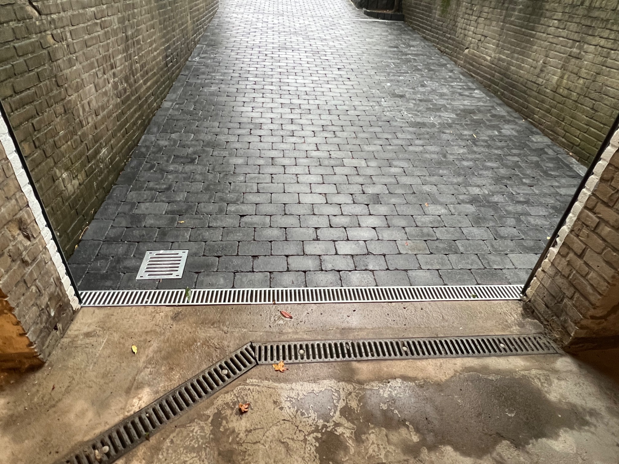 Ułożona kostka brukowa w wąskim przejściu między budynkami z systemem odprowadzania wody, widoczne kratki ściekowe i spad na nawierzchni.