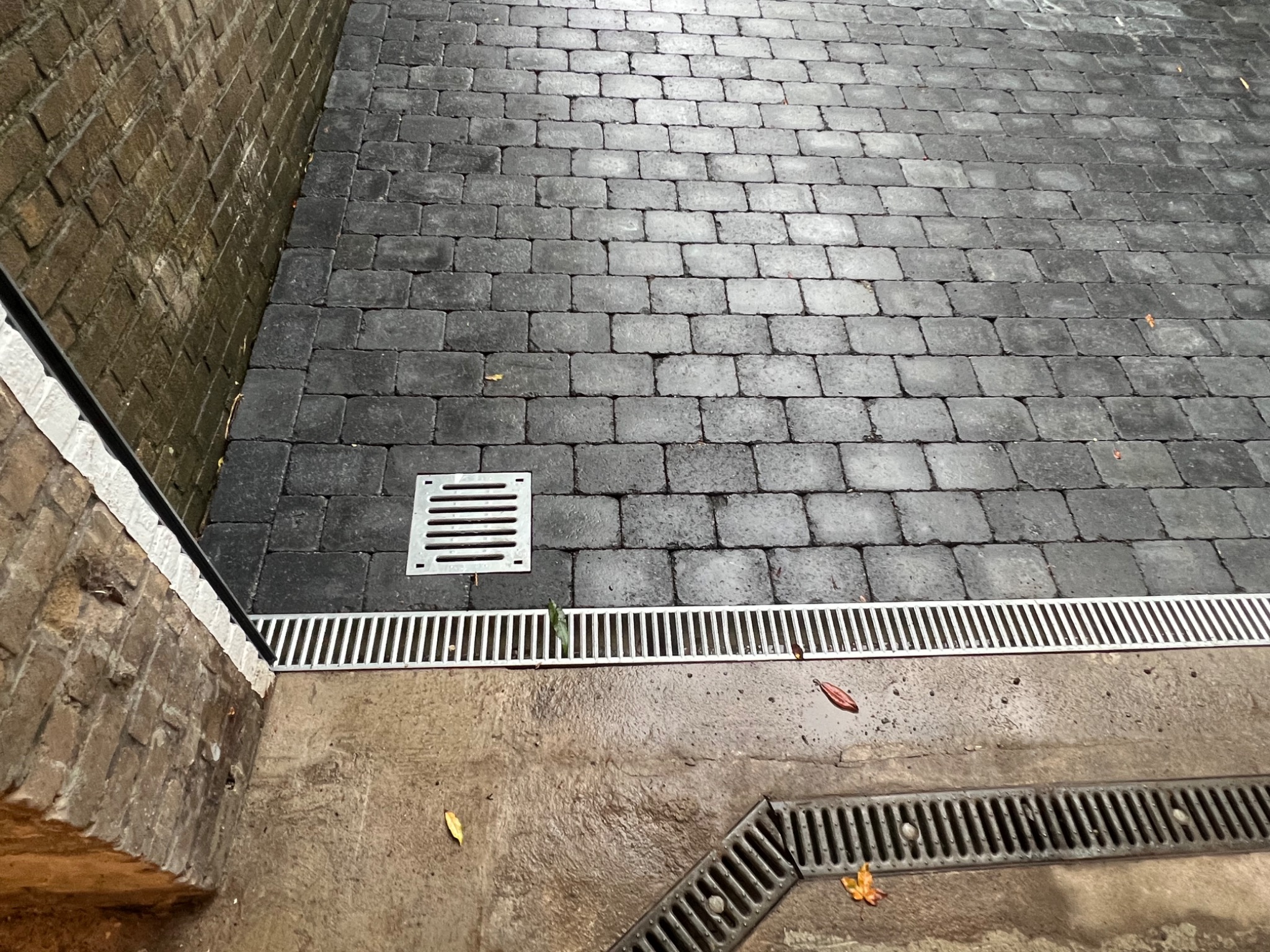 Brukowana nawierzchnia z odpływem liniowym i kratką ściekową, widoczne liście na mokrej powierzchni.