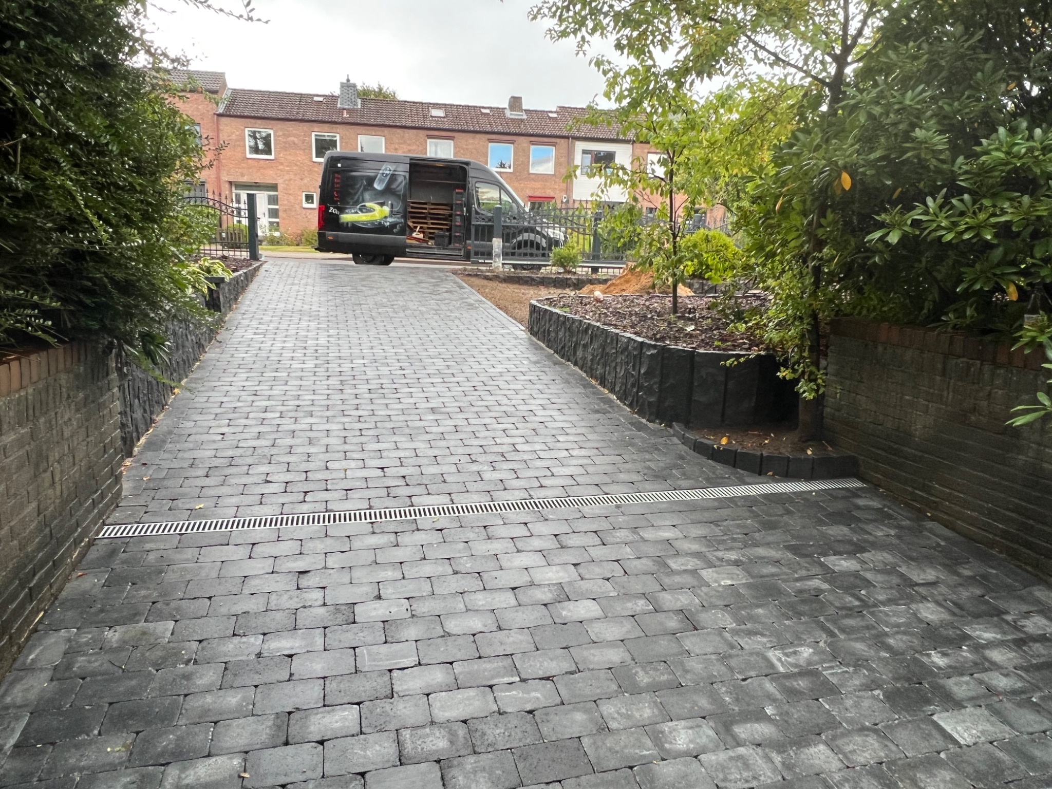 Ułożona kostka brukowa na podjeździe, z liniowym odpływem wody, murki po bokach, samochód dostawczy w tle.