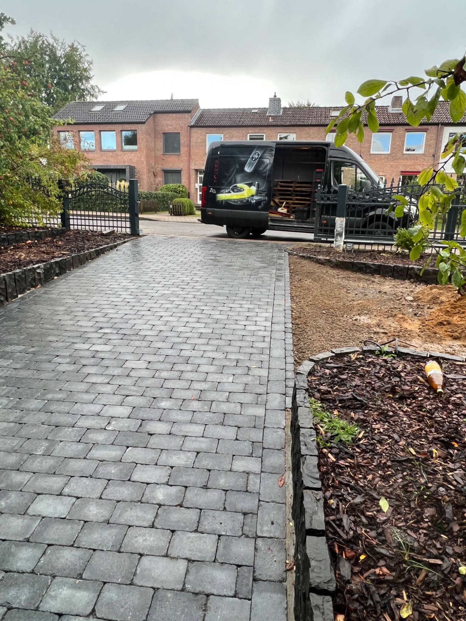 Ułożona, szara kostka brukowa prowadząca do bramy wjazdowej, obok częściowo przygotowane podłoże pod rabatę i otwarty samochód dostawczy firmy w tle.