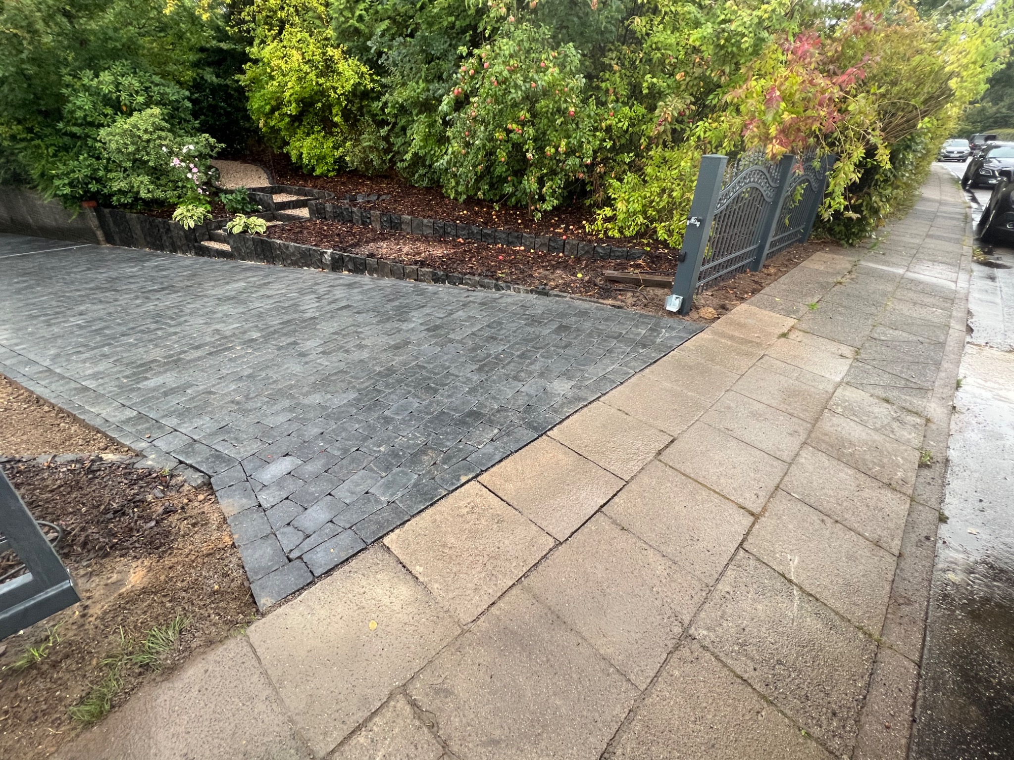 Ułożona kostka brukowa w kolorze grafitowym, oddzielona od chodnika z jasnych płyt betonowych, metalowe ogrodzenie w tle i roślinność wzdłuż granicy posesji.