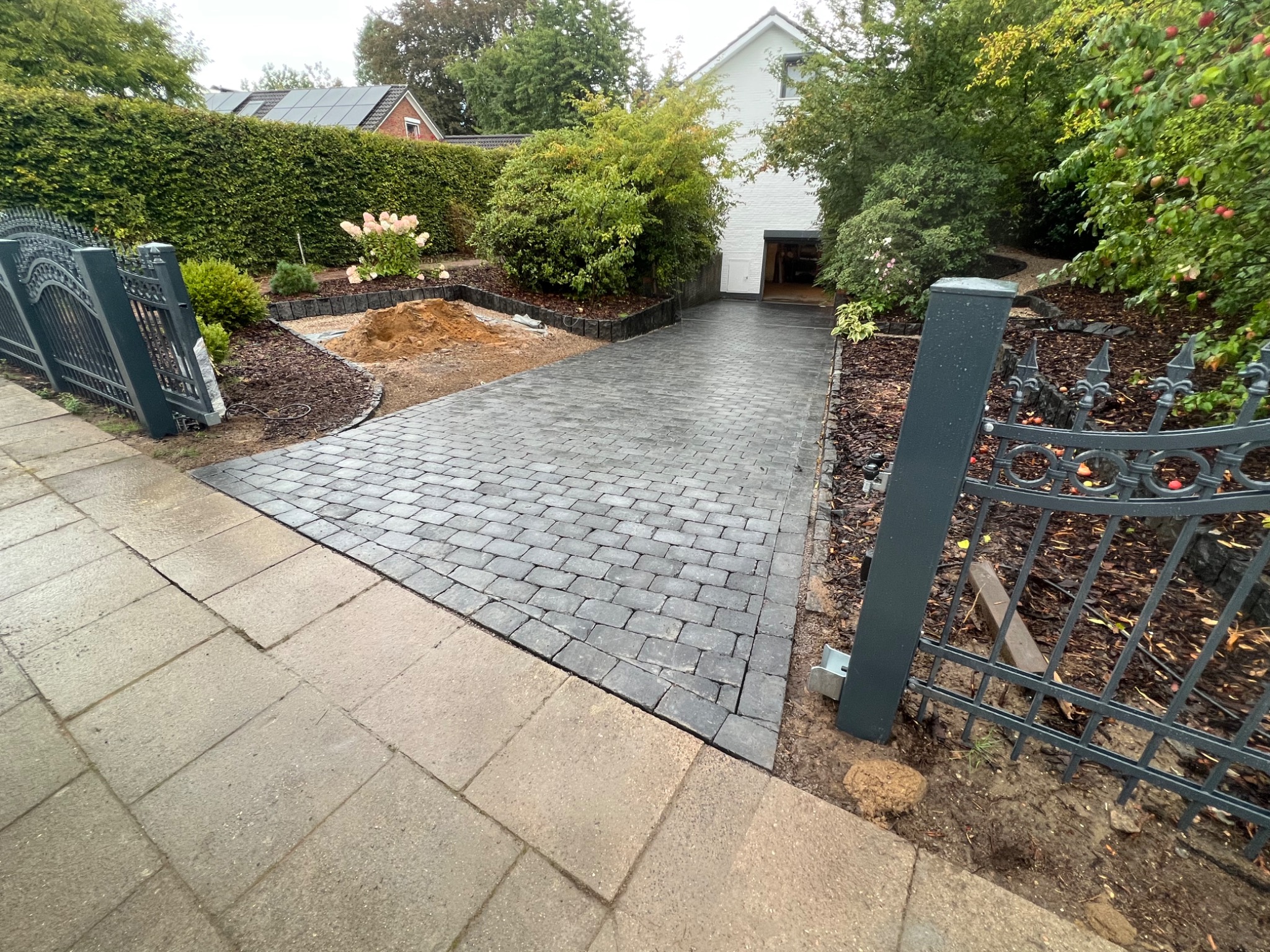 Ułożona kostka brukowa w kolorze grafitowym prowadząca do garażu, widoczne rabaty z korą, częściowo otwarte metalowe ogrodzenie i trawnik z tujami w tle.