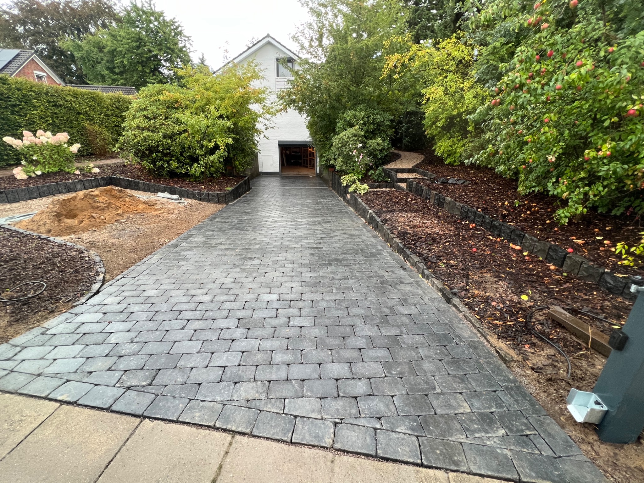 Ułożona kostka brukowa prowadząca do garażu, obok rabaty z korą i roślinnością, częściowo przygotowany teren pod nasadzenia po lewej stronie.