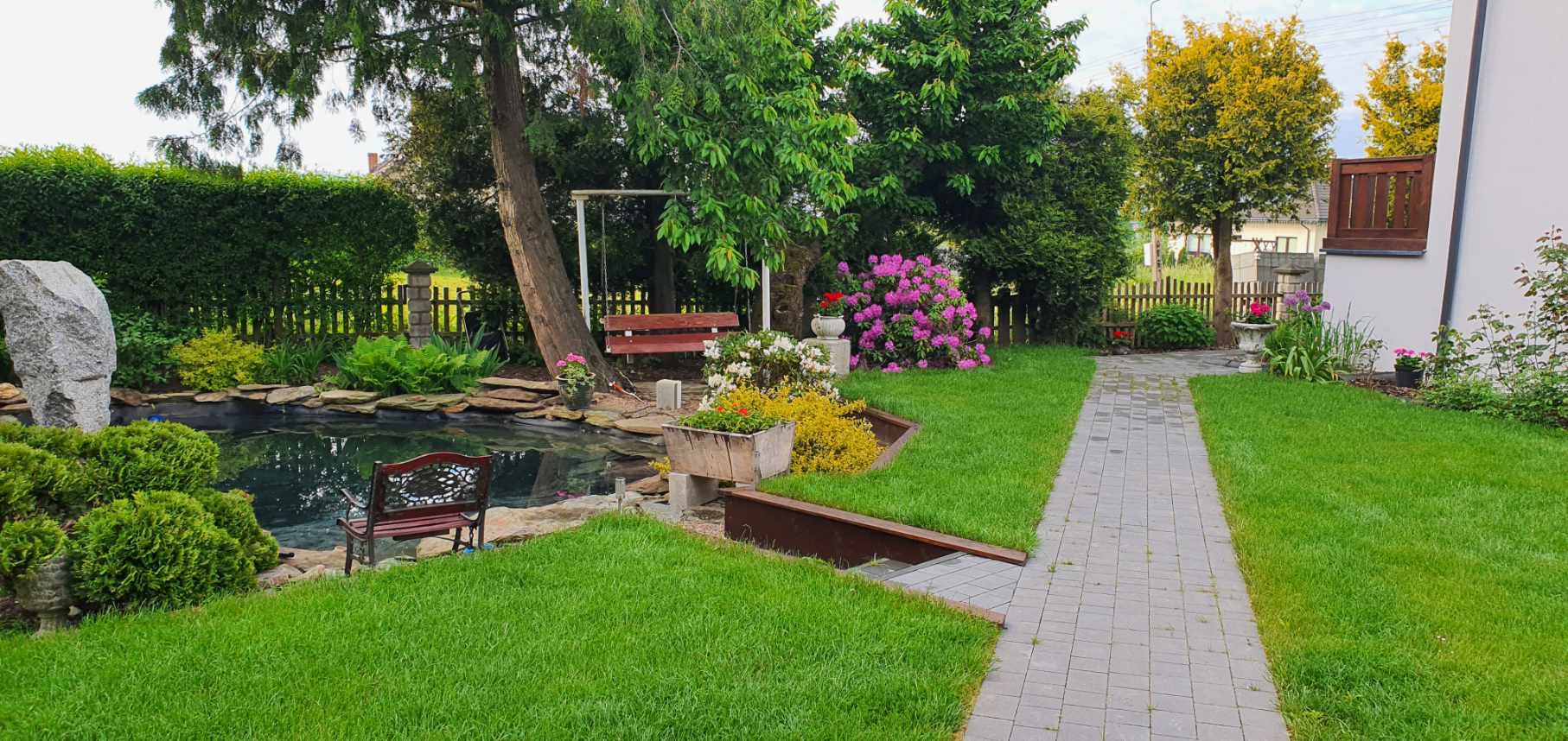 Malowniczy ogród z oczkiem wodnym, huśtawką-ławka pod drzewem i kamienną ścieżką, otoczony bujną zielenią i kwitnącymi krzewami.