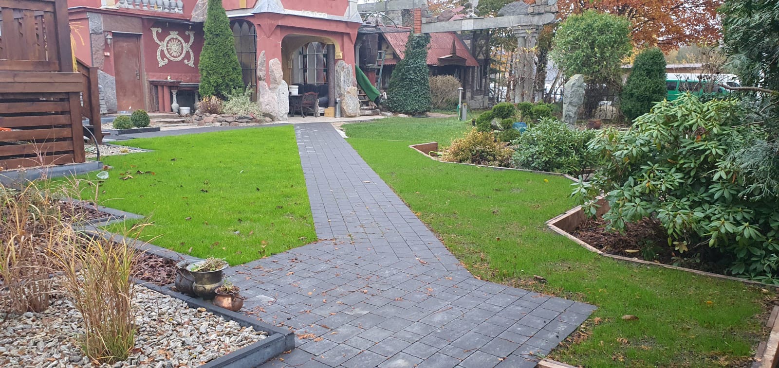 Widok na zadbany ogród z równo przystrzyżonym trawnikiem, ścieżką z kostki brukowej prowadzącą do nietypowego budynku z elementami dekoracyjnymi, drzewami i krzewami w tle. Widać fragment systemu...