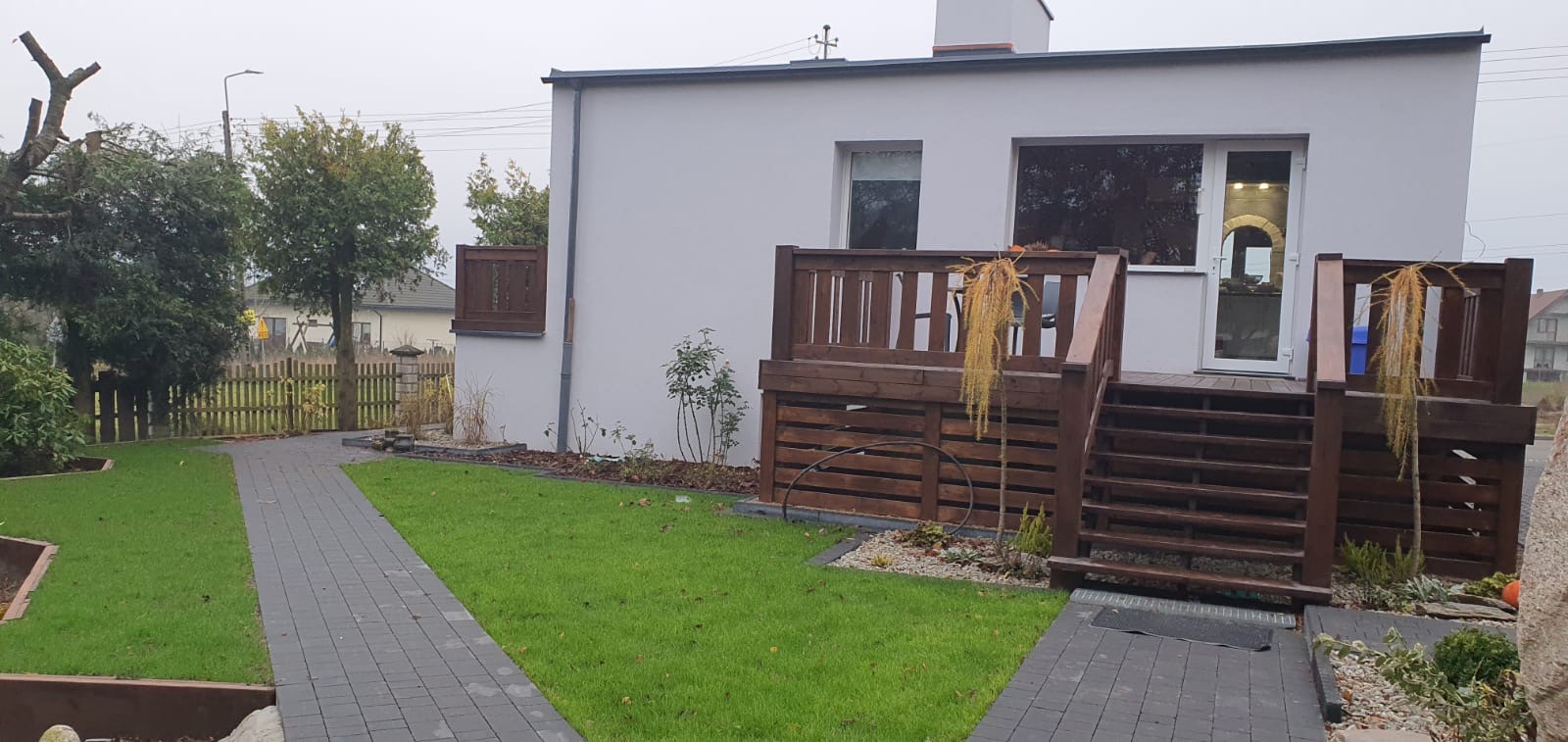 Nowoczesny dom z drewnianym tarasem i schodami, widok na zadbany trawnik i ścieżkę z kostki brukowej.