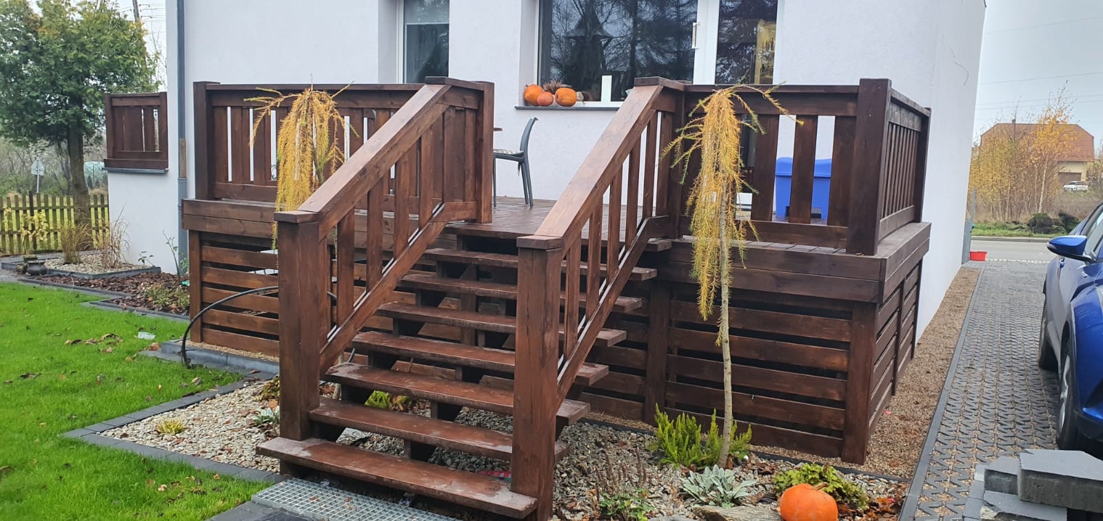 Podwyższony taras z drewna z schodami, otoczony ozdobnymi roślinami i dyniami, widok na dom z białymi ścianami i szarą kostką brukową.