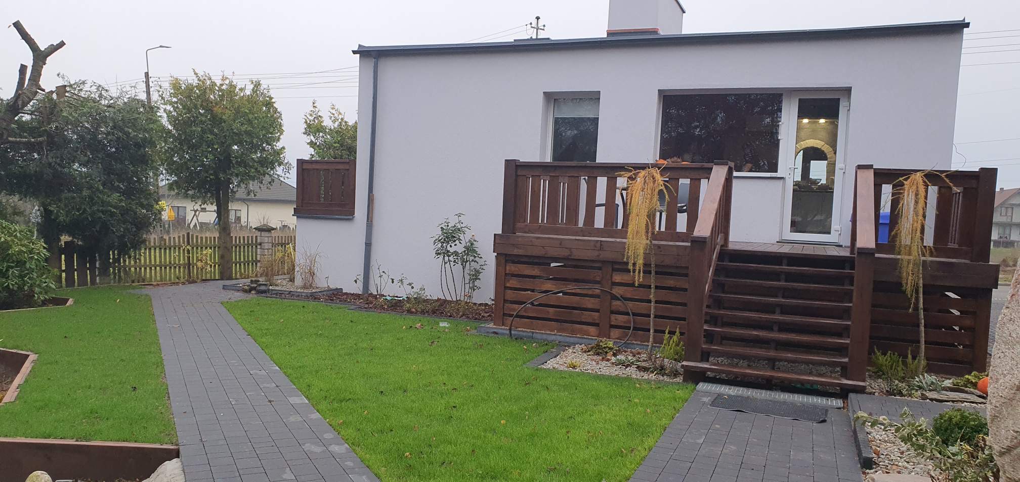 Nowoczesny dom z tarasem drewnianym z barierką, schodami i małym drzewkiem iglastym, trawnik z kostką brukową, widok z zewnątrz w pochmurny dzień.