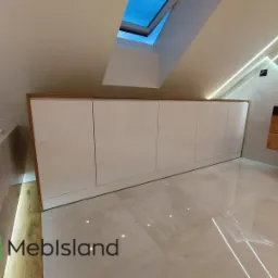 Biała szafa wnękowa pod skosem dachu w łazience z oknem dachowym, widoczne oświetlenie LED i logo firmy w rogu.