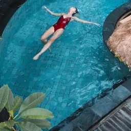 Kobieta w czerwonym kostiumie kąpielowym unosi się na plecach w basenie z turkusową wodą i ciemną, kamienną obudową, otoczonym tropikalną roślinnością, widziana z góry.
