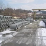 Stos szarych, betonowych rur ułożonych w rzędach na placu składowym. W tle budynki przemysłowe i żuraw. Fragmenty śniegu na ziemi.
