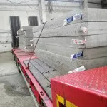 Ciężarówka z czerwonym nadwoziem przewozi palety szarych, betonowych płyt, zabezpieczonych niebieską folią. Widoczne oświetlenie magazynowe w tle.